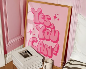 Può includere: Un poster incorniciato rosa con il testo "Yes, You Can!" in uno stile di font retrò. Il poster è appeso a una parete rosa e in primo piano c'è una pila di libri e una tazza su un pavimento di legno.