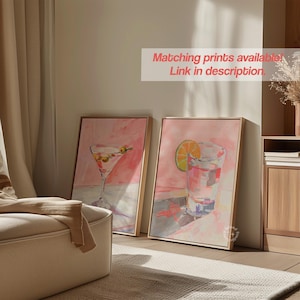 May include: Two framed watercolor paintings of cocktails. One depicts a martini with olives, the other a drink with a lime slice. The paintings are in shades of pink and gold, with the text "Matching prints available! Link in description."