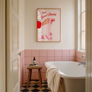 May include: A bathroom with pink tile walls and a white bathtub. A framed print with the text "Hello Sweet Cheeks" hangs on the wall above the bathtub. A wooden stool with a bottle of hand soap sits next to the bathtub.
