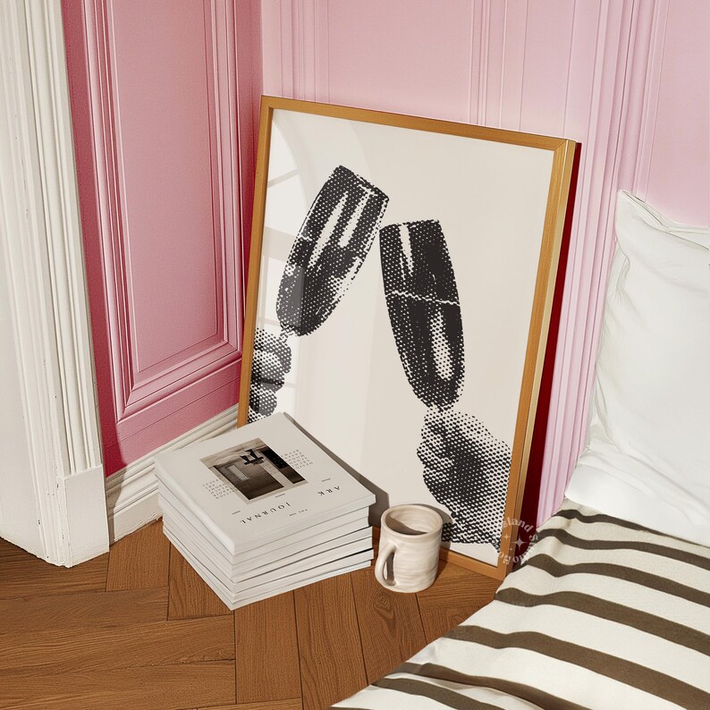 May include: A framed black and white print of two champagne glasses clinking together. The print is in a wooden frame and is hanging on a pink wall.