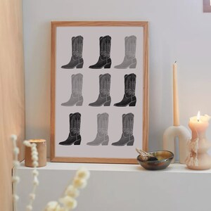 May include: A framed print of nine black and white cowboy boots in a gradient pattern. The boots are arranged in three rows of three. The print is framed in a light wood frame.