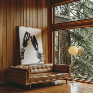 May include: A brown leather sofa with tufted upholstery sits in a mid-century modern living room with wood paneling and a large window overlooking a lush green forest. A black and white print of two glasses clinking hangs on the wall above the sofa. A floor lamp with a round white shade illuminates the room.