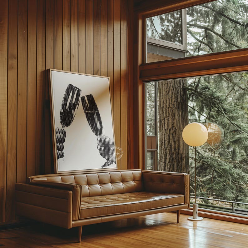 May include: A brown leather sofa with tufted upholstery sits in a mid-century modern living room with wood paneling and a large window overlooking a lush green forest. A black and white print of two glasses clinking hangs on the wall above the sofa. A floor lamp with a round white shade illuminates the room.