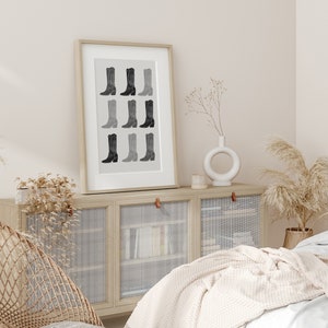 May include: A framed black and white print of a row of cowboy boots in various shades of gray. The print is hanging on a wall above a light wood cabinet with glass doors.
