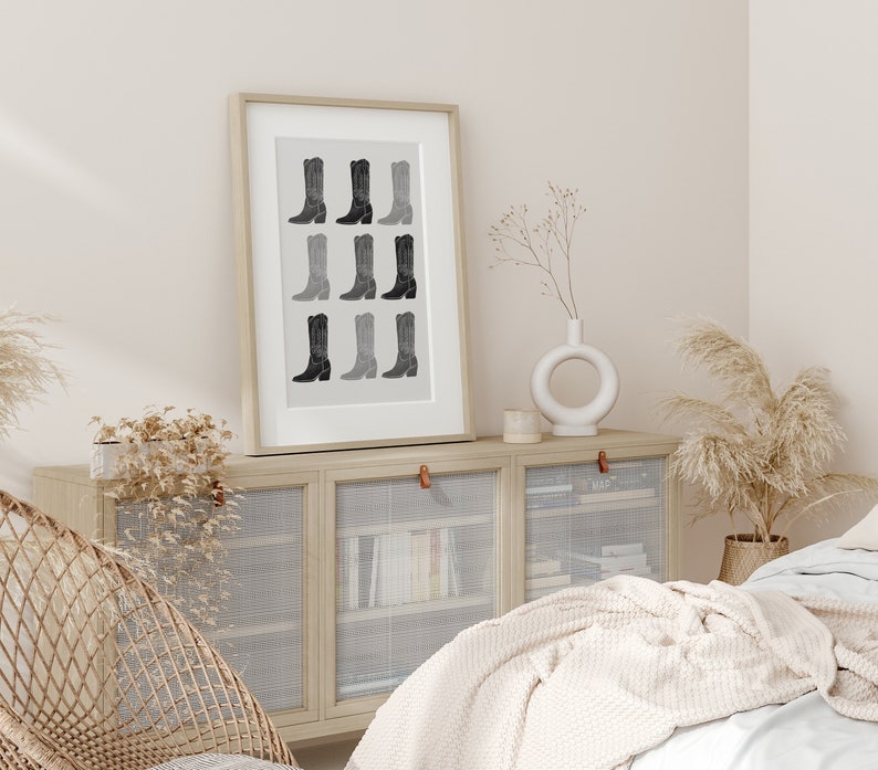 May include: A framed black and white print of a row of cowboy boots in various shades of gray. The print is hanging on a wall above a light wood cabinet with glass doors.