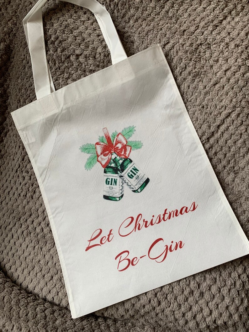 Pode incluir: Sacola de tecido branca com um la&ccedil;o vermelho e duas garrafas verdes que dizem "Gin". O texto "Let Christmas Be-Gin" est&aacute; impresso em vermelho na sacola.