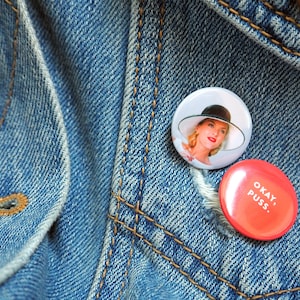 Op de afbeelding: Twee knoopsluitingen op een blauw denim jack. Eén knoopsluiting heeft een vrouw met een witte hoed en een witte jurk. De andere knoopsluiting is rood met witte tekst die "Okay, Puss." zegt.