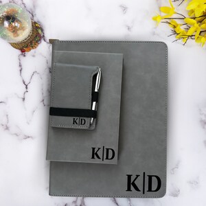 K&ouml;nnte beinhalten: Ein Set aus drei grauen Leder-Notizb&uuml;chern mit einem schwarzen Gummiband und einem Stifthalter. Die Notizb&uuml;cher sind mit den Initialen "K|D" personalisiert.