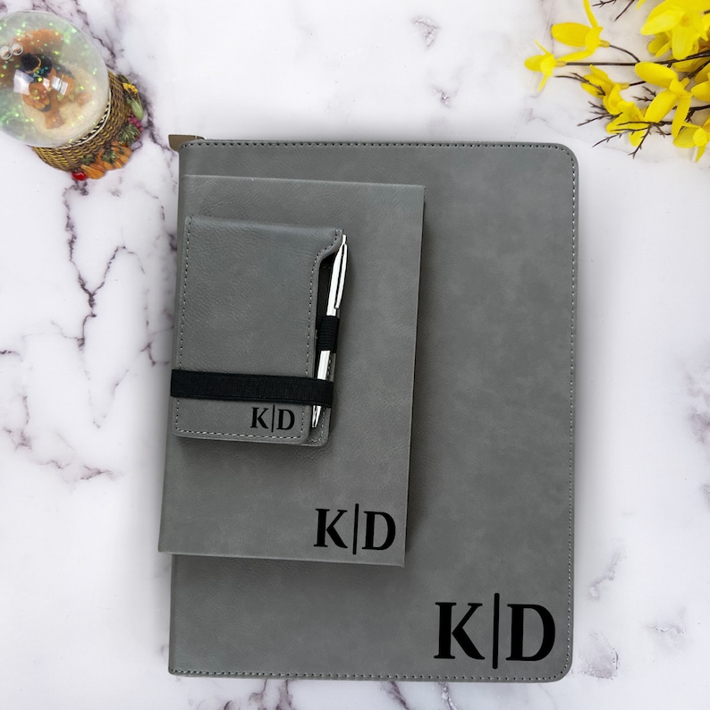 K&ouml;nnte beinhalten: Ein Set aus drei grauen Leder-Notizb&uuml;chern mit einem schwarzen Gummiband und einem Stifthalter. Die Notizb&uuml;cher sind mit den Initialen "K|D" personalisiert.
