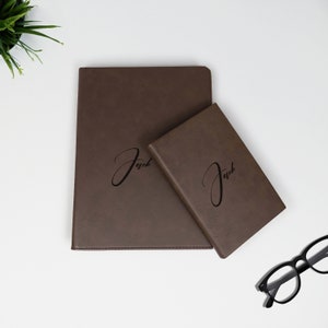 K&ouml;nnte beinhalten: Zwei braune Lederjournale mit dem Namen "Jack" auf der Vorderseite gepr&auml;gt. Das gr&ouml;&szlig;ere Journal befindet sich unten und das kleinere Journal oben.