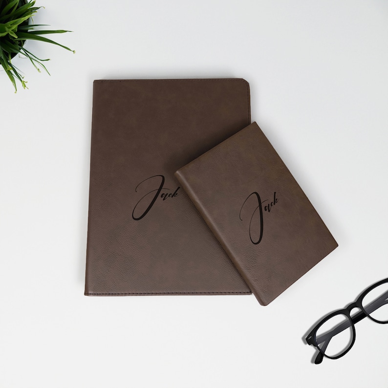 K&ouml;nnte beinhalten: Zwei braune Lederjournale mit dem Namen "Jack" auf der Vorderseite gepr&auml;gt. Das gr&ouml;&szlig;ere Journal befindet sich unten und das kleinere Journal oben.