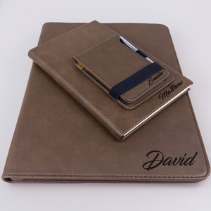 May include: Three brown leather journals with personalized names: David, Emilia, and Matthew. The journals are stacked on top of each other, with the largest journal on the bottom and the smallest journal on top.