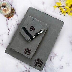 K&ouml;nnte beinhalten: Drei graue Lederjournale mit einem schwarzen Gummibandverschluss und einem personalisierten Monogramm in einem Kreis. Das Monogramm ist "GMB".