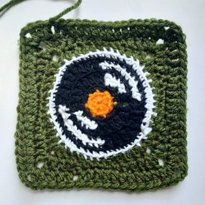 Puede incluir: Un cuadrado de crochet verde con un círculo de crochet negro y blanco en el centro. El círculo tiene un pequeño círculo naranja en el centro.