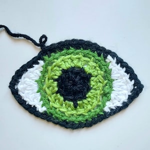 Peut inclure: Un œil crocheté avec un iris vert, une pupille noire et une sclère blanche. L'œil est entouré d'une bordure noire.