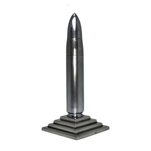 Puede incluir: Un tapón de botella plateado con forma de bala. El tapón tiene una punta y un cuerpo cilíndrico. Está montado sobre una base cuadrada escalonada. El objeto es de metal y tiene una superficie reflectante.