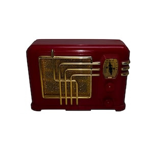 May include: A retro-style radio in a deep red finish. The radio has gold-coloured accents, including a speaker grill and decorative trim. The dial and knobs are also gold, adding to the vintage aesthetic. The radio is a classic design.