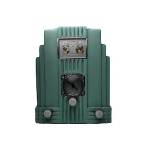 Puede incluir: Un radio reloj de estilo art déco de color verde azulado. El reloj tiene una esfera plateada y dos perillas. Encima de la esfera hay un pequeño panel rectangular con dos globos. El radio reloj mide aproximadamente 20 cm de alto.