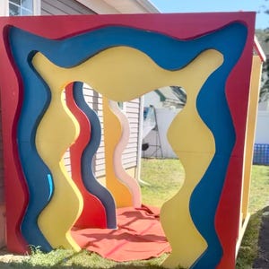 May include: A colorful, abstract play structure with a red, blue, and yellow design. The structure has wavy cutouts and is set on a grassy lawn. The primary colors are red, blue, and yellow.