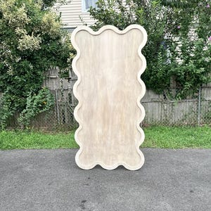 May include: A large, light-colored wooden wall mirror with a unique wavy, scalloped edge. The mirror is rectangular and stands upright on a paved surface, with a natural wood grain finish. The background includes greenery and a house.