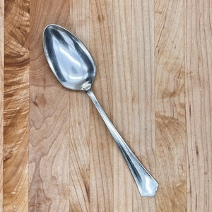 Puede incluir: Una cuchara de plata con un cuenco redondeado y un mango decorativo. La cuchara está sobre una superficie de madera.