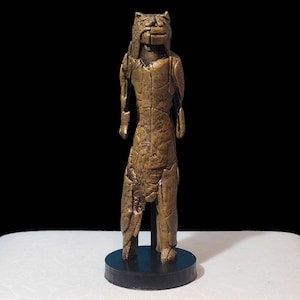 Puede incluir: Una escultura de color bronce de una figura de pie con cabeza felina, posiblemente un jaguar o animal similar. La figura tiene una superficie texturizada y está montada sobre una base circular negra.