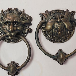 Puede incluir: Dos llamadores de puerta ornamentados con un diseño de rostro grotesco. Los llamadores están hechos de metal y tienen un acabado bronce oscuro. Los rostros están detallados con una superficie texturizada y tienen una expresión amenazante.