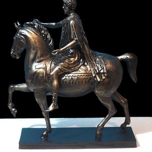 Op de afbeelding: Een bronzen beeld van een ruiter te paard. De ruiter draagt een toga en houdt de teugels in zijn linkerhand. Het paard staat op een zwarte sokkel.