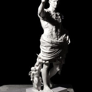 May include: White marble statue of a Roman emperor standing with his right arm extended, pointing forward. He is wearing a toga and a breastplate with intricate details. A smaller statue of a winged figure is at his feet.
