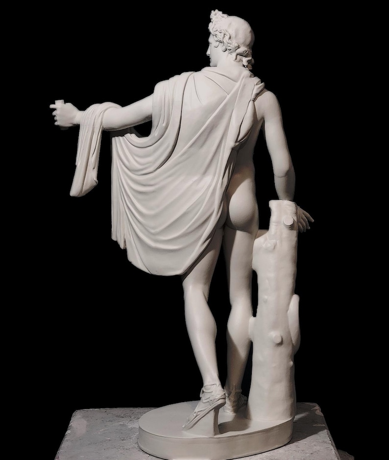 May include: A white plaster sculpture of a nude male figure standing with a drapery draped over his left arm. The figure is holding a small object in his right hand and is leaning against a tree trunk.