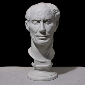 Puede incluir: Busto de yeso blanco de un hombre con expresión seria. La escultura representa a un hombre con pelo corto, nariz prominente y mandíbula definida. El busto está montado sobre una base de pedestal y el fondo es negro.
