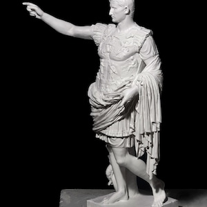 May include: White marble statue of a Roman emperor, standing with his right arm extended, pointing with his index finger. He is wearing a toga and a breastplate with intricate details. The statue is on a white square base.