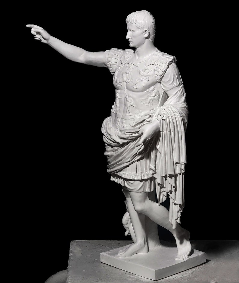 May include: White marble statue of a Roman emperor, standing with his right arm extended, pointing with his index finger. He is wearing a toga and a breastplate with intricate details. The statue is on a white square base.
