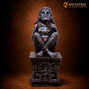 May include: A dark bronze-toned sculpture of a seated figure with an expressive face, looking upward with hands raised near the chest. The figure features intricate carvings on the limbs and torso, resting on a square pedestal with geometric patterns.