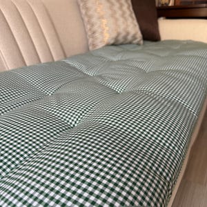 Puede incluir: Un sofá beige con un cojín a cuadros verdes y blancos. Dos cojines decorativos en tonos neutros descansan contra el respaldo del sofá. El cojín tiene un diseño acolchado, creando un aspecto clásico y cómodo.