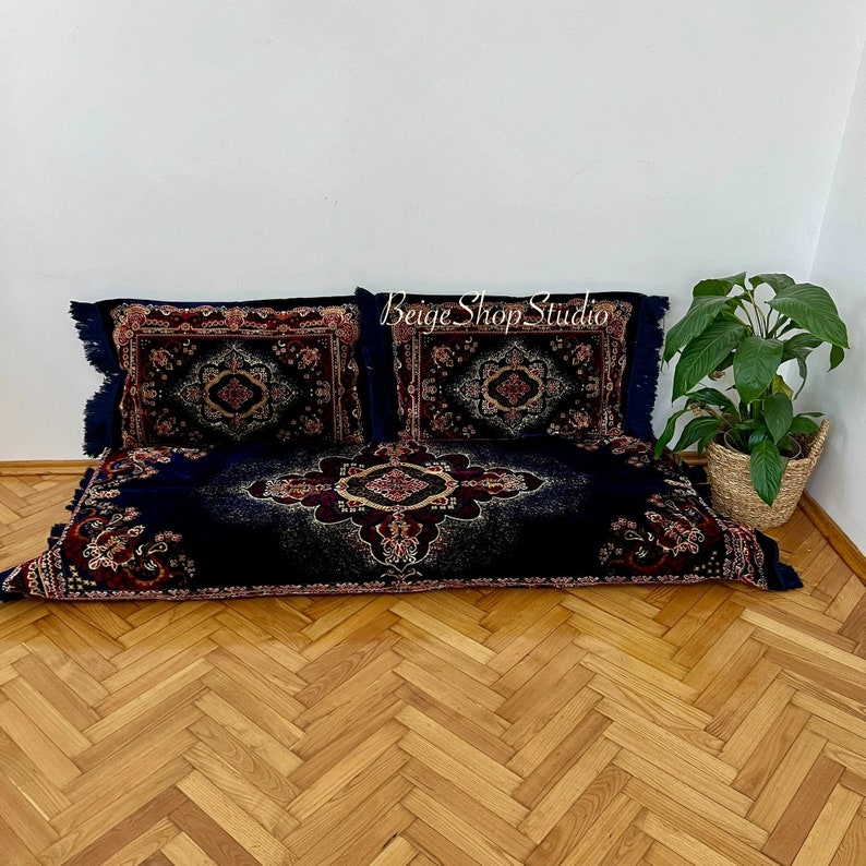 May include: Two blue floor cushions with intricate red, gold, and black patterned designs. The cushions have fringe trim and are arranged on a wood floor.