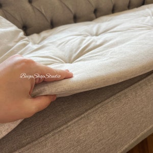 May include: A close-up of a light gray sofa cushion with a hand lifting the edge to reveal the underside of the cushion. The cushion is tufted and has a soft, plush texture.