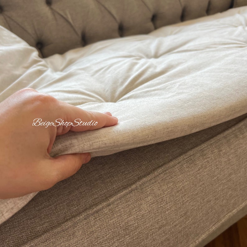 May include: A close-up of a light gray sofa cushion with a hand lifting the edge to reveal the underside of the cushion. The cushion is tufted and has a soft, plush texture.