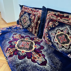 May include: Two blue throw pillows with intricate red, gold, and white patterns on a blue velvet sofa with a similar pattern. The pillows have fringe trim.