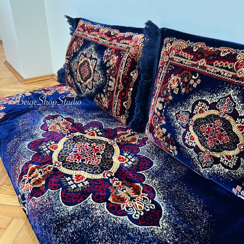 May include: Two blue throw pillows with intricate red, gold, and white patterns on a blue velvet sofa with a similar pattern. The pillows have fringe trim.