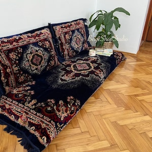 May include: A navy blue floor cushion with a red, gold, and black patterned design. The cushion is decorated with fringe and has two matching pillows.