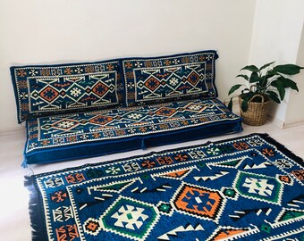 Può includere: Un set di cuscini da pavimento con motivo blu e arancione con un design geometrico. Il set comprende due cuscini grandi e un tappeto più piccolo. I cuscini sono disposti in fila su un pavimento bianco.