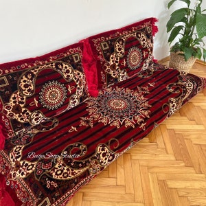 May include: Red and gold patterned floor cushions with fringe. The cushions are arranged in a row on a wooden floor. The cushions are decorated with intricate floral designs.