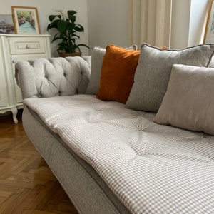 May include: A light gray sofa with a brown and white checkered cushion. The sofa has a tufted back and is decorated with several throw pillows, including a burnt orange pillow.