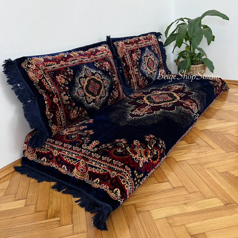 May include: A navy blue floor cushion with a red, gold, and white patterned design. The cushion has fringe trim and is topped with two matching pillows.