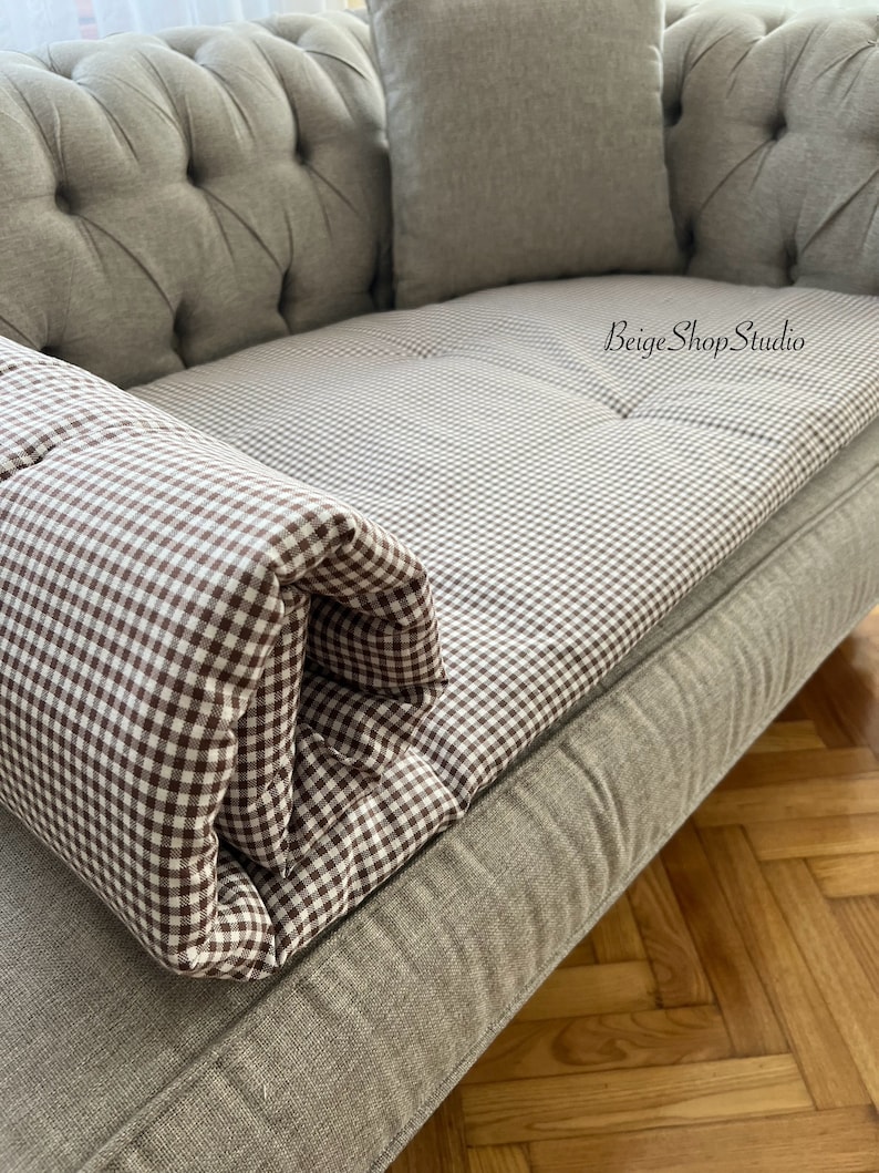 May include: A gray tufted sofa with a brown and white gingham patterned cushion. The cushion is rolled up and placed on the arm of the sofa. The sofa is in a room with a wooden floor.