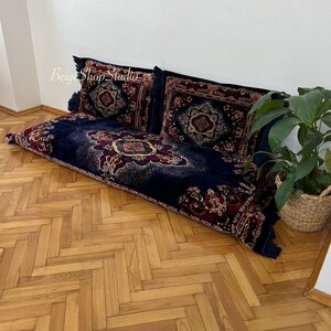 May include: A navy blue floor cushion with a red, gold, and white patterned design. The cushion is made of velvet and has a fringe trim. The cushion is sitting on a wooden floor with a herringbone pattern.