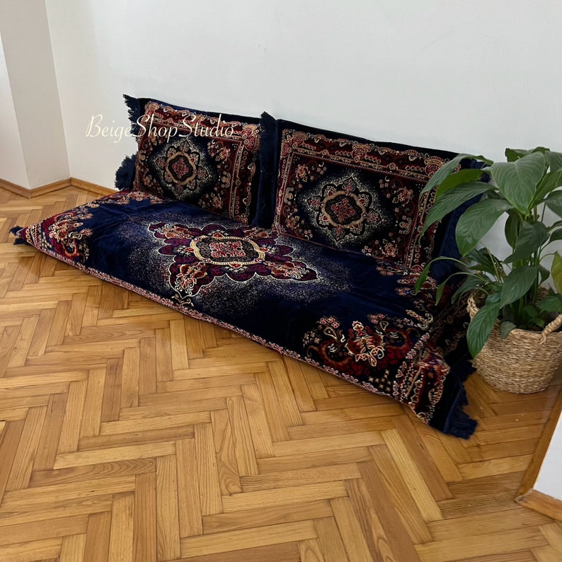 May include: A navy blue floor cushion with a red, gold, and white patterned design. The cushion is made of velvet and has a fringe trim. The cushion is sitting on a wooden floor with a herringbone pattern.