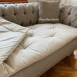 May include: A light gray tufted sofa with a white throw pillow and a white throw blanket. The sofa has a wooden frame and legs. The text "Beige Shop Studio" is visible on the sofa.