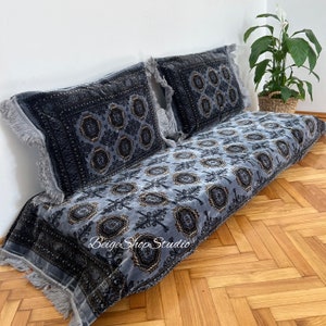 Puede incluir: Un cojín de suelo gris y negro con estampado con dos almohadas a juego. El cojín tiene un borde de flecos y las almohadas tienen borlas. El cojín está en un suelo de madera.